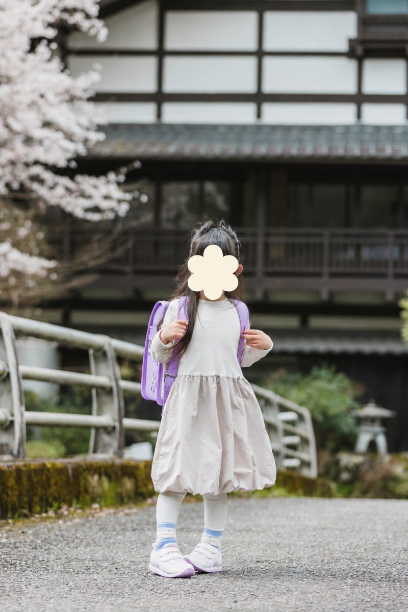 ランドセルを背負って立つ娘。後ろに桜と建物が見える