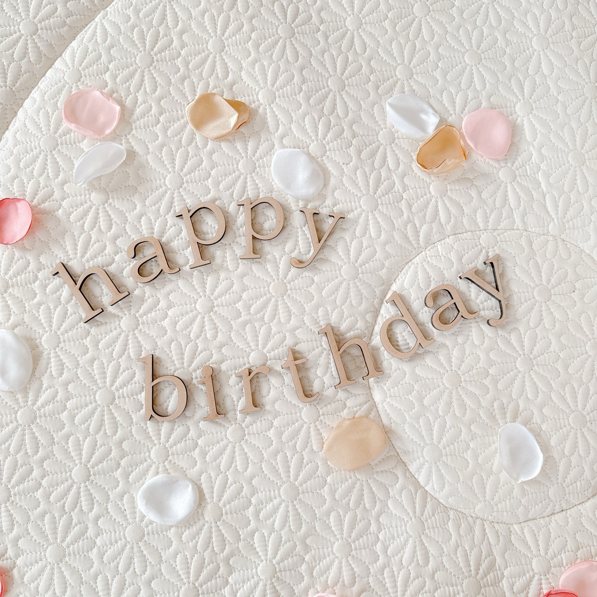 「happy birthday」の木製レターバナーをホワイトキルトの上に並べた床置きスタイル。花びらと一緒に置くだけで記念日感が出ます