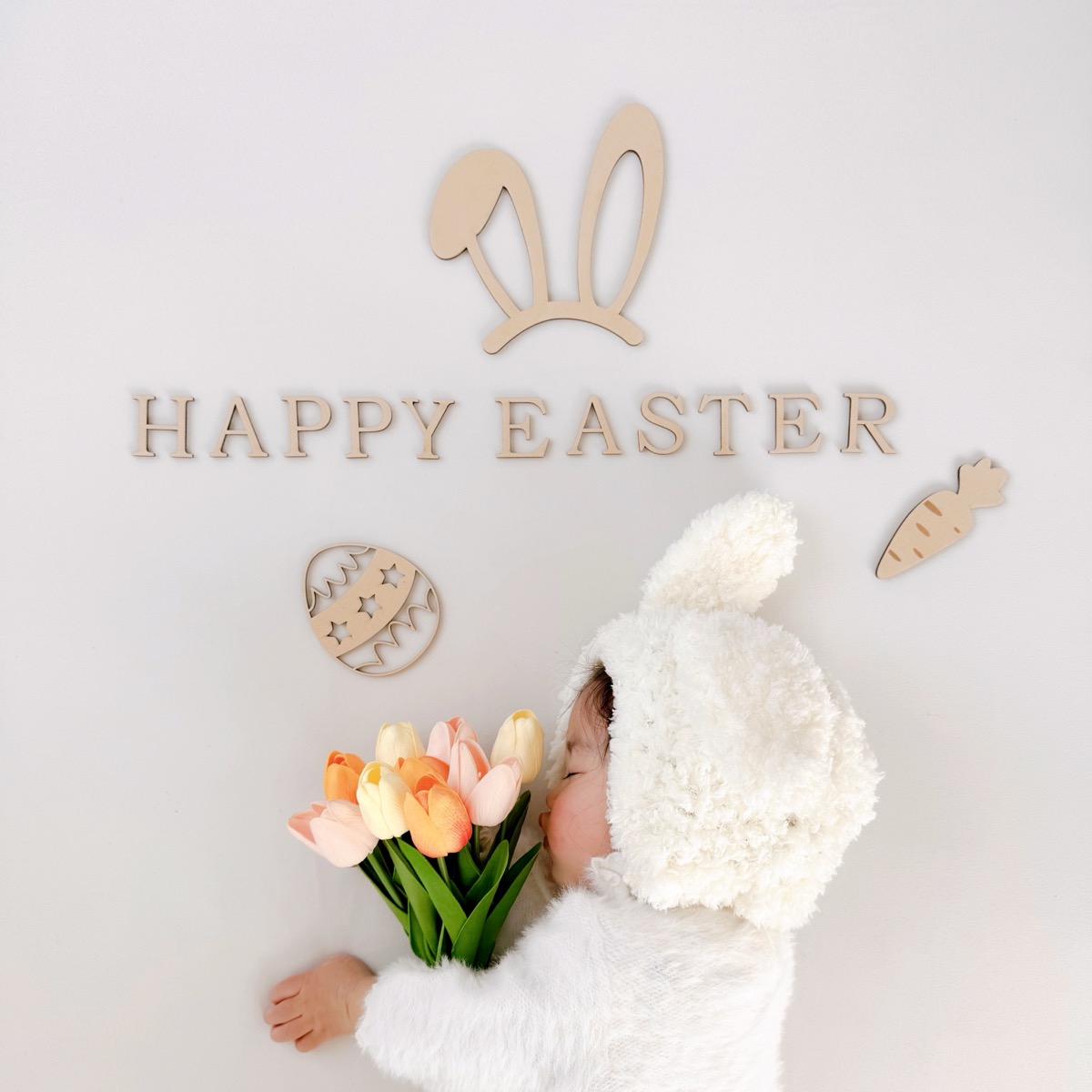 白い壁に木製の「HAPPY EASTER」バナーを貼り、バニー衣装の赤ちゃんを前に座らせた壁貼りスタイルの撮影例