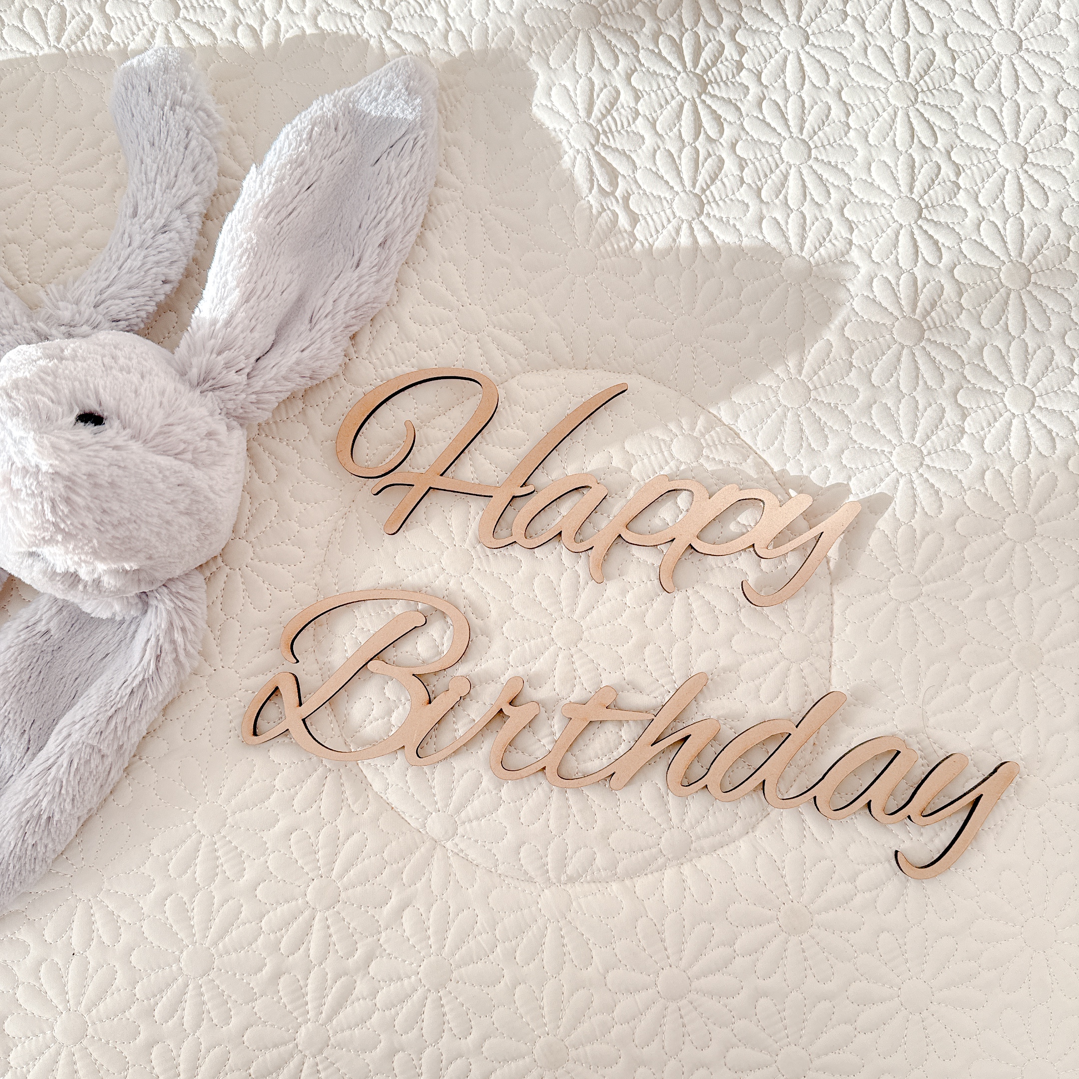 Happy Birthdayの木製レターバナーとうさぎのぬいぐるみ。白いキルトの上に並べたやさしい雰囲気の誕生日小物