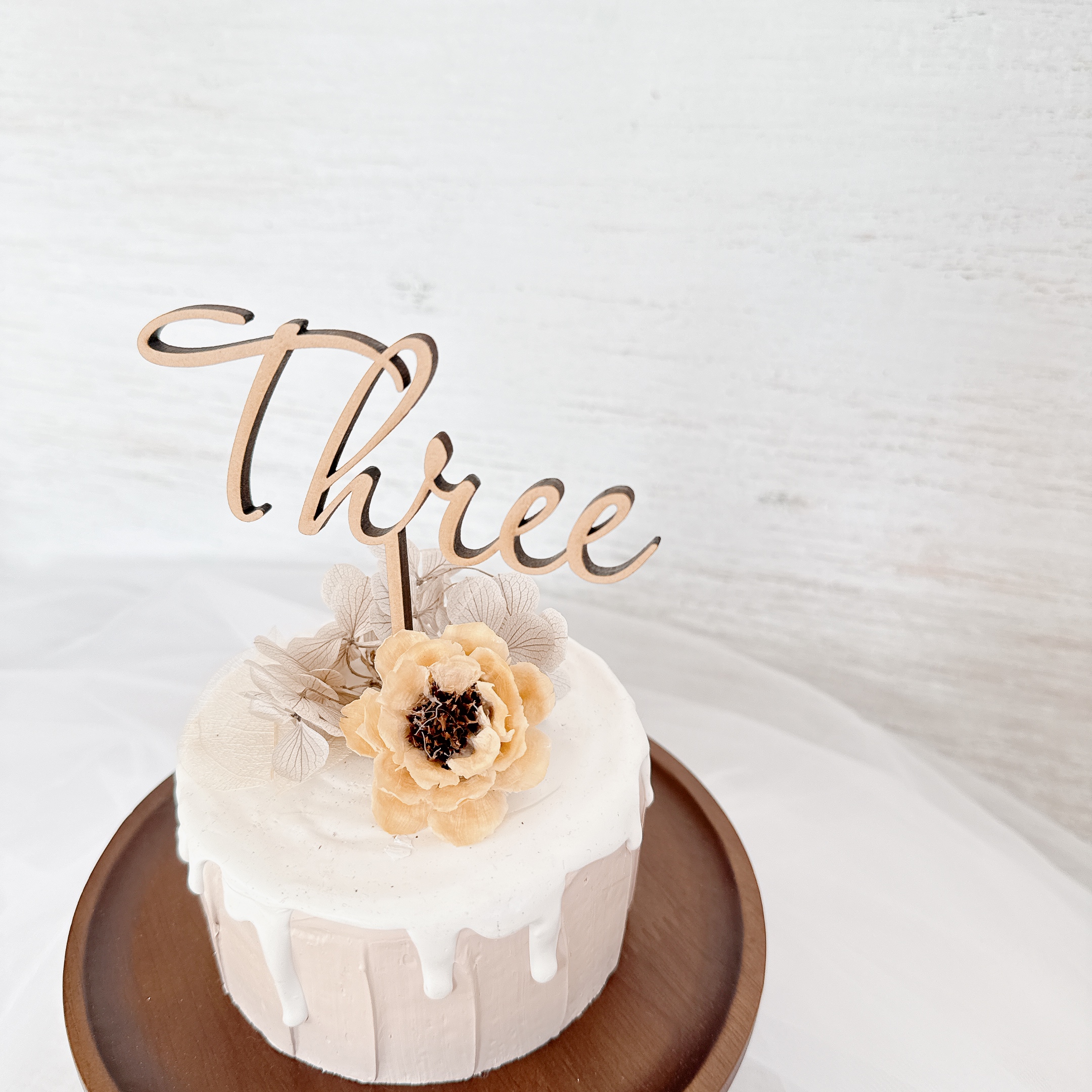 「Three」の木製ケーキトッパーを飾ったホワイトケーキ。3歳の誕生日にぴったりのシンプルでおしゃれな一台